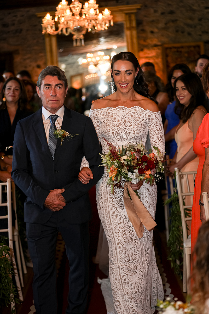 The bride walks down the aisle with her father to meet her groom, holding her wedding bouquet which is beautifully designed with red, orange, green and white florals.