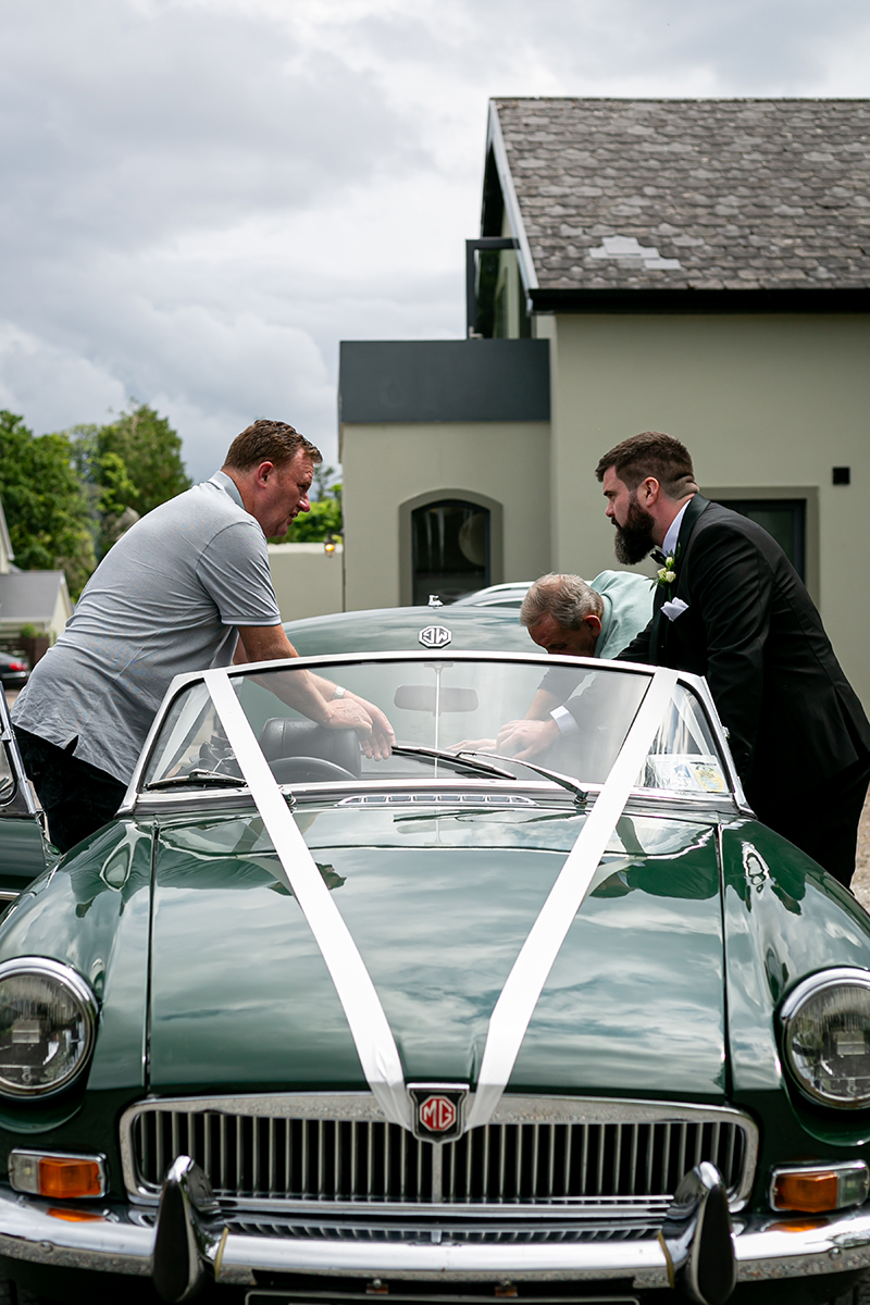 Three groomsmen check on the wedding car, a dark green cabriolet.