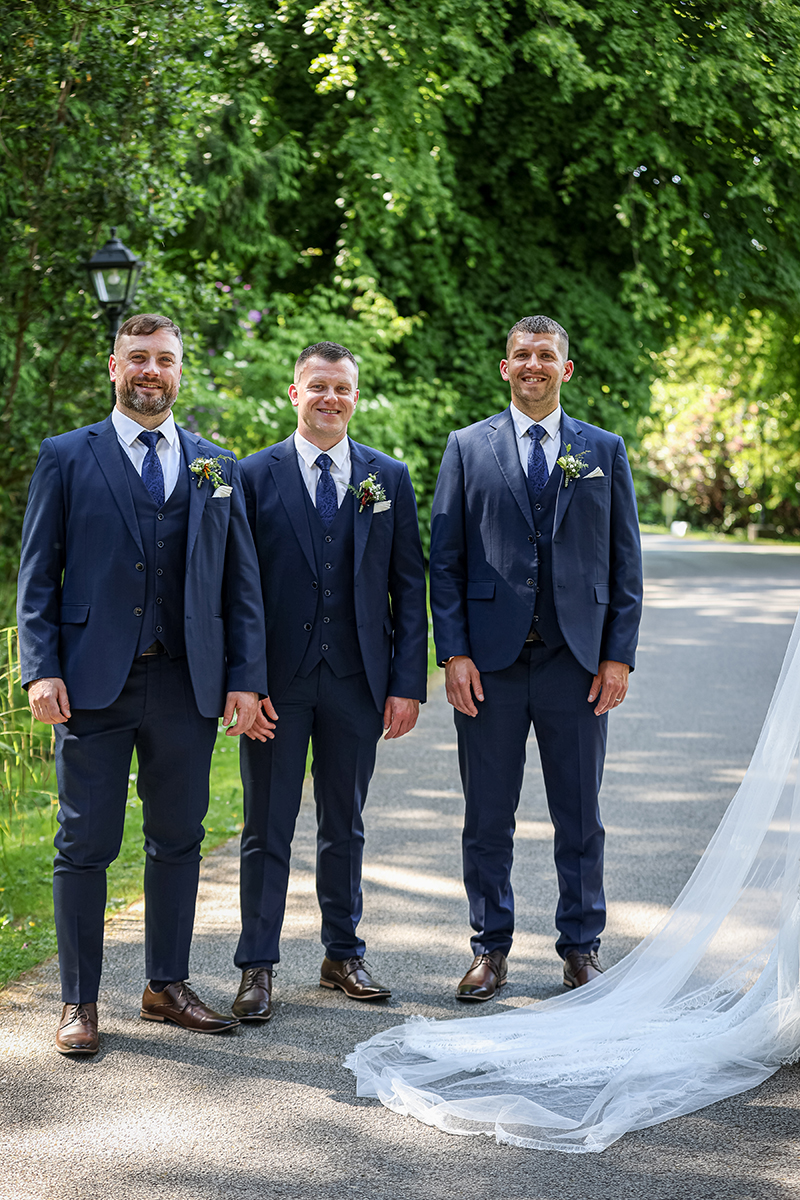 Groomsmen stand in the grounds of Ballyseede castle surrounded by green foliage and sunshine.