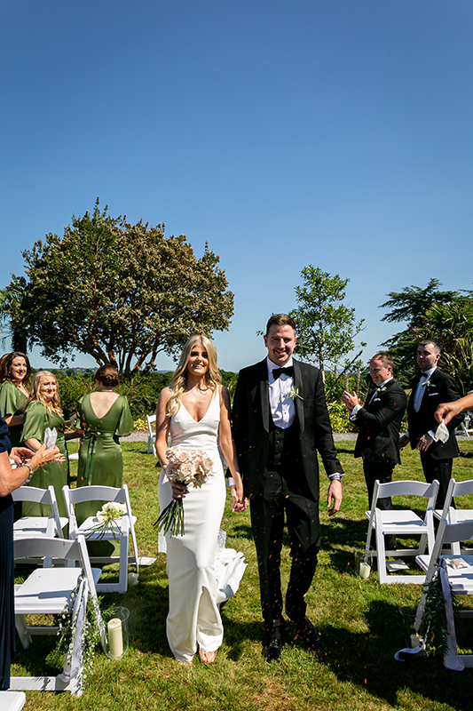 bride and groom walk down the aisle at the dunloe hotel gounds hand in hand with the bride holding her bouquet in her right hand
