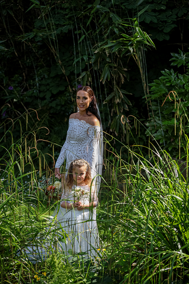 The bride and her daughter stand amongst green shrubs smiling at the camera on the grounds of Ballyseede castle hotel, Tralee.
