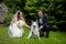 Bride and groom pictured kneeling in the green grass with a dog sitting in between them, surrounded by foliage of the Dromquinna manor grounds.