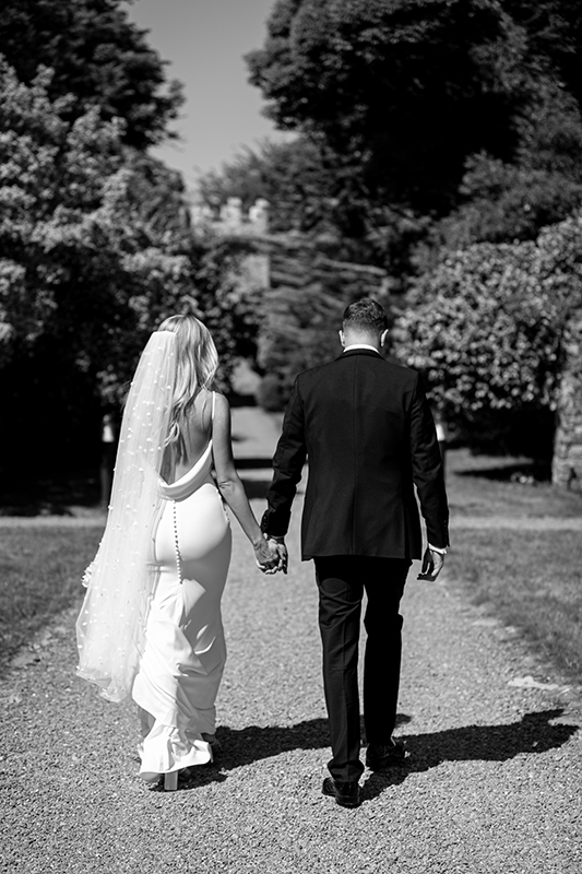 black and white portrait of bride and groom holding hands walking away from camera along a path near the dunloe hotel
