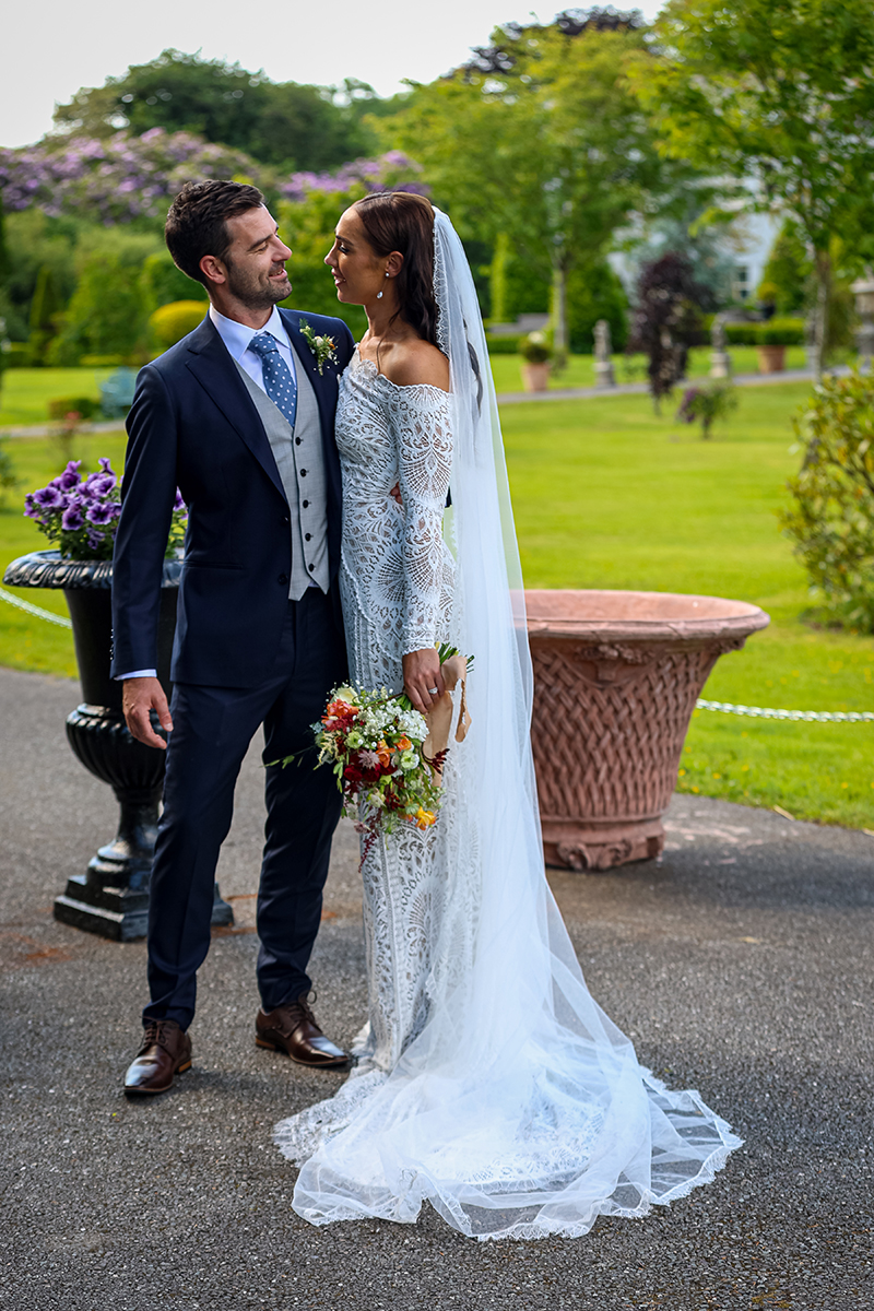 Bride and groom stand gazing at each other lovingly with the soft green gardens of Ballyseede castle hotel in the background.