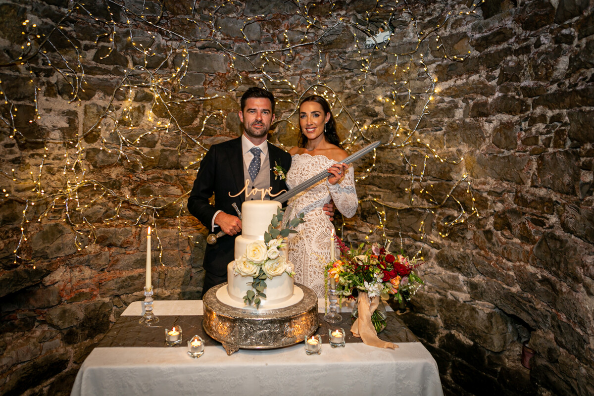 Bride and groom pose behind their wedding cake holding a ceremonial sword to cut the cake at their wedding reception in Ballyseede castle, Tralee.