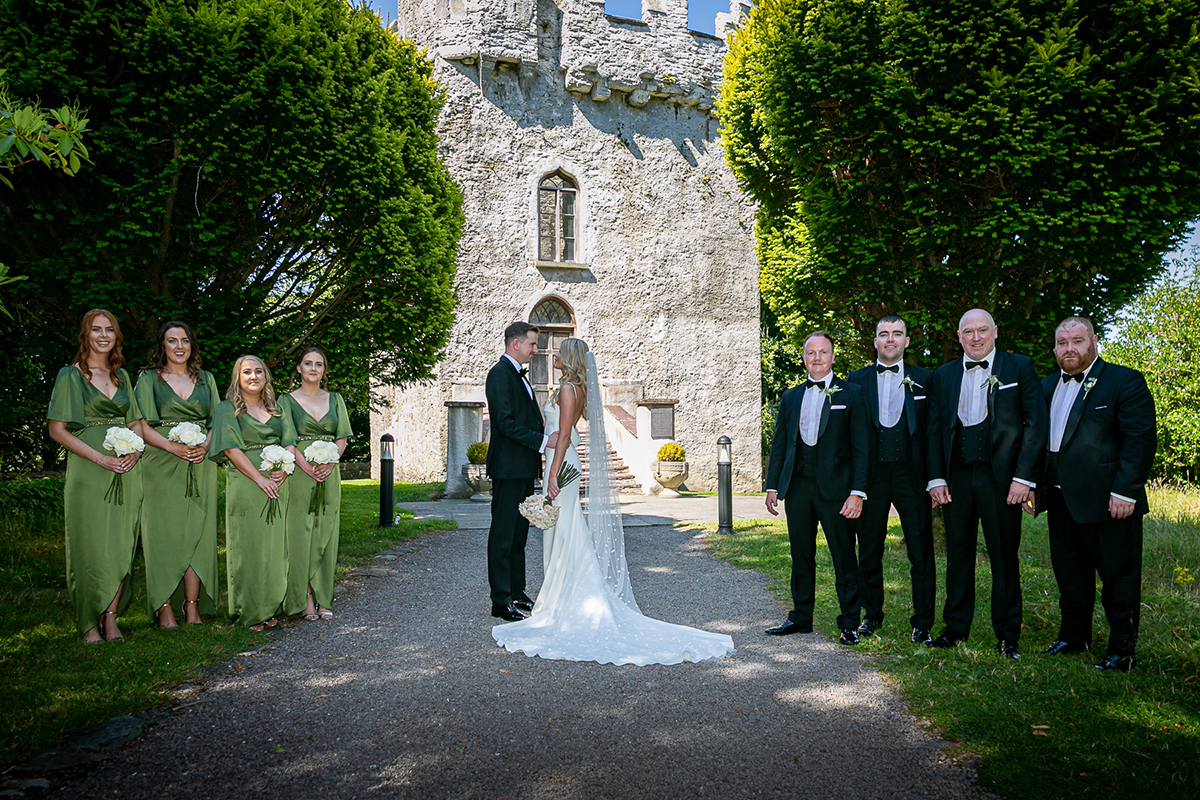 bride and groom are stood centred facing each other with the bridesmaids to the left side and the groomsmen to the right with a beautiful castle in the background