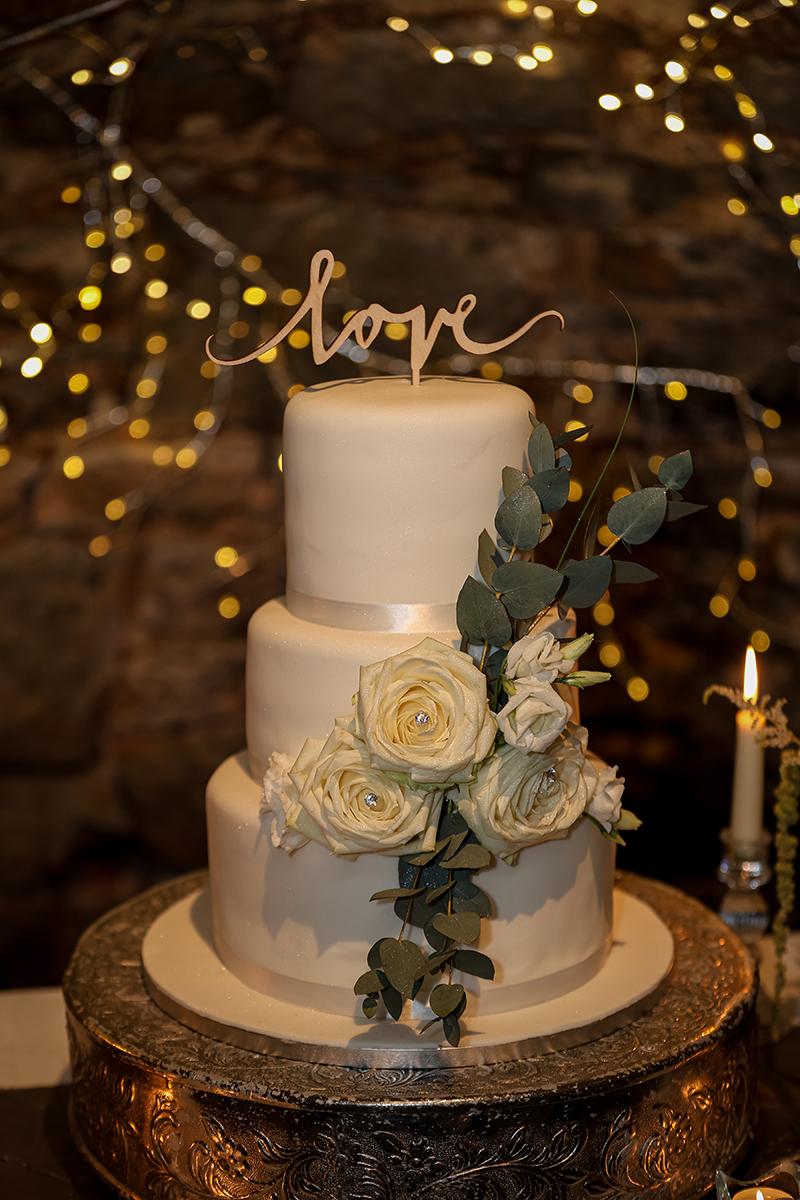Portrait of the wedding cake dressed in white roses and greenery with soft bokeh twinkly lights in background at Ballyseede castle, Tralee.
