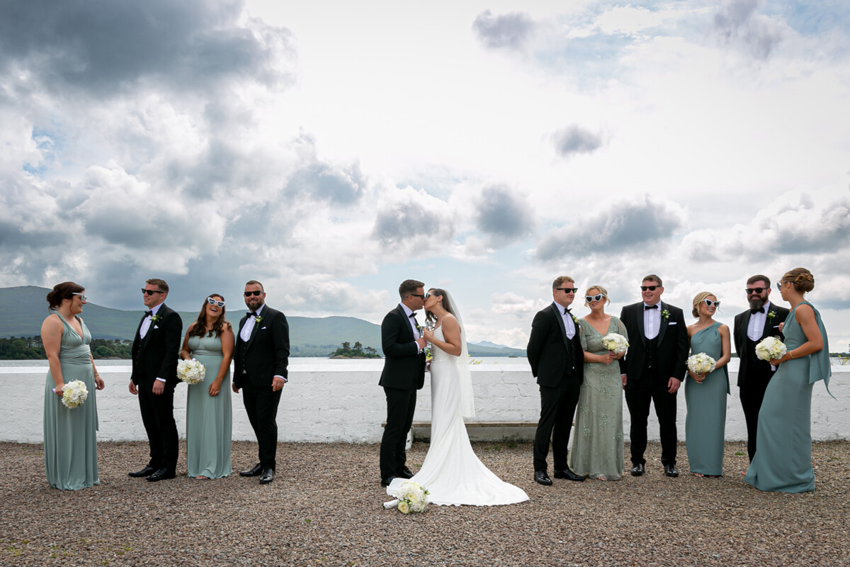 Bride and groom are pictured sharing a kiss with their wedding party stood on either side of them, with the beautiful mountains of Kenmare and blue skies in the background.