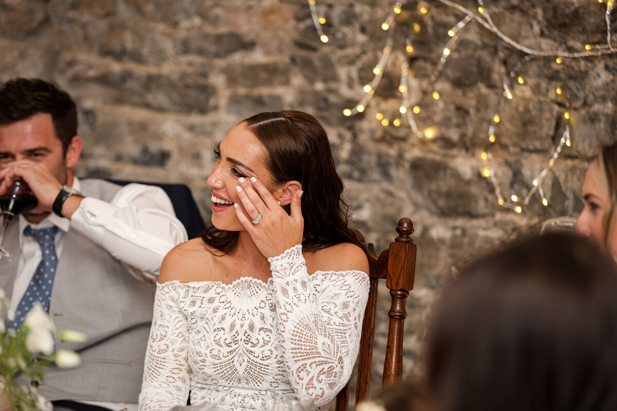 The bride wipes a tear of happiness from her eyes while laughing during the wedding reception at Ballyseede castle hotel, with a soft stone background and bokeh lights.