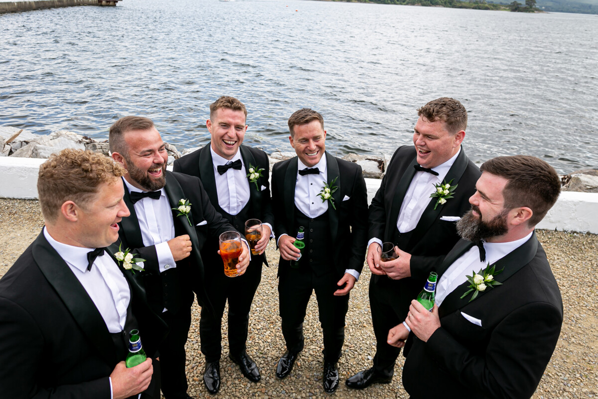 Groom and his groomsmen stand in a circle smiling and enjoying a drink with soft waves of Kenmare bay in the background.