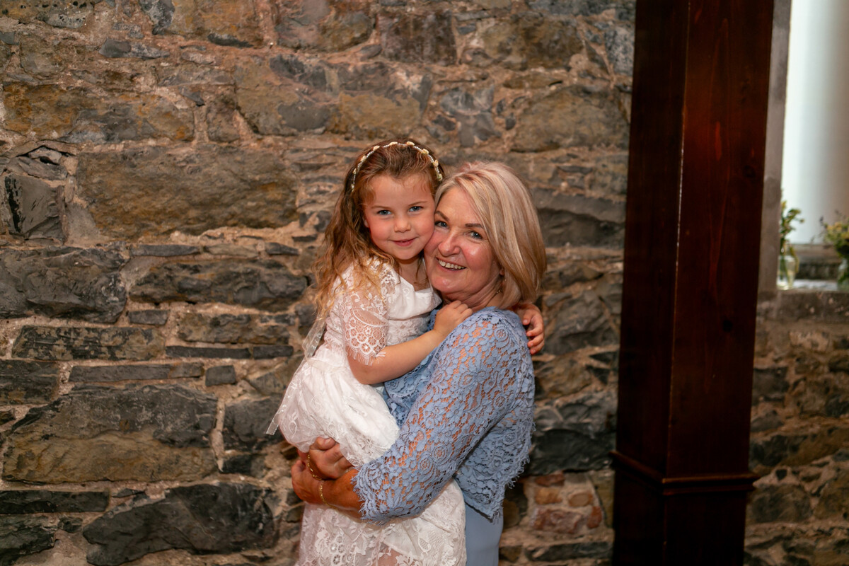 Grandmother with granddaughter stand in front of a brick wall smiling at the camera enjoying the wedding at Ballyseede castle in Tralee.