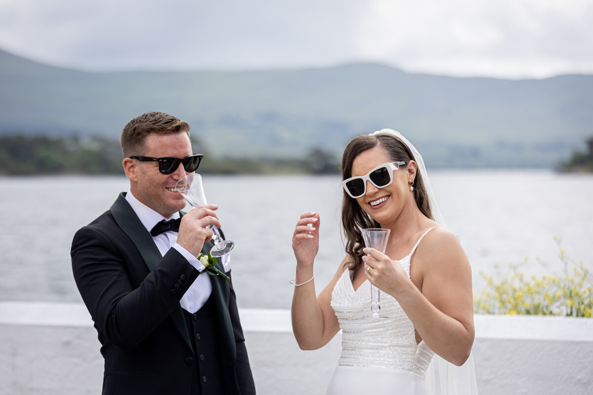 The bride and groom are pictured wearing sunglasses and enjoying a glass of champagne with a soft blurred background of Kenmare bay and surrounding mountain scape.