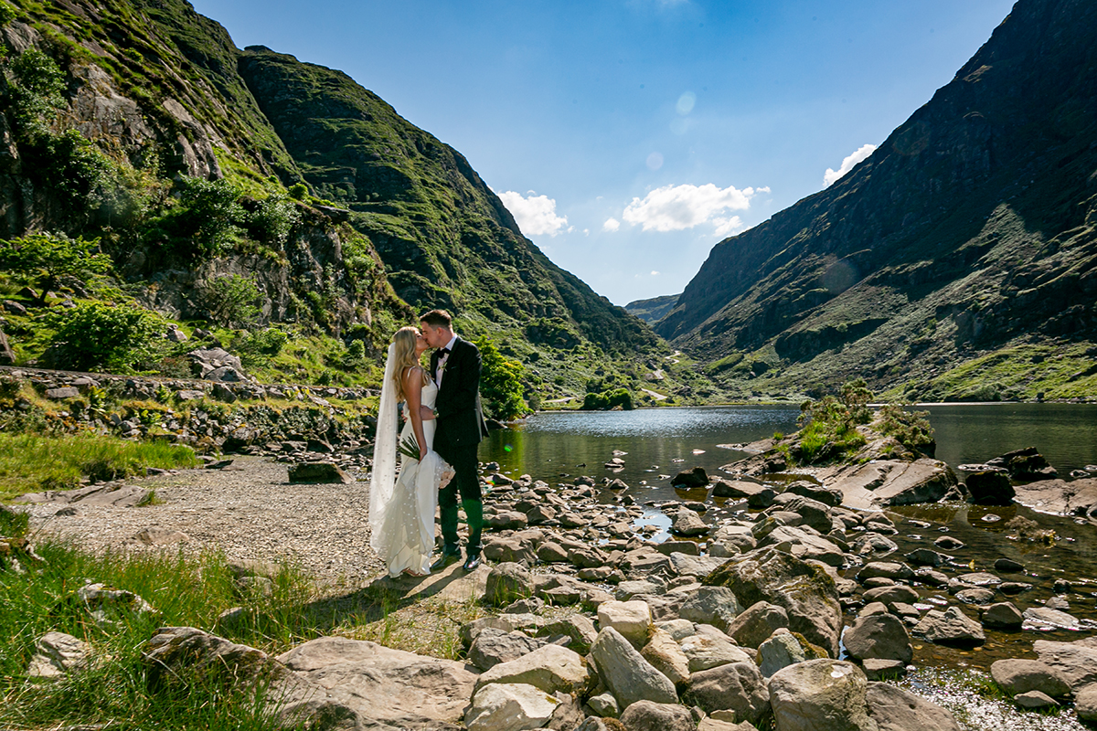 panoramic image of the gap of dunloe with the bride and groom embracing and sharing a kiss standing by the water surrounded bu mountains and blue skies