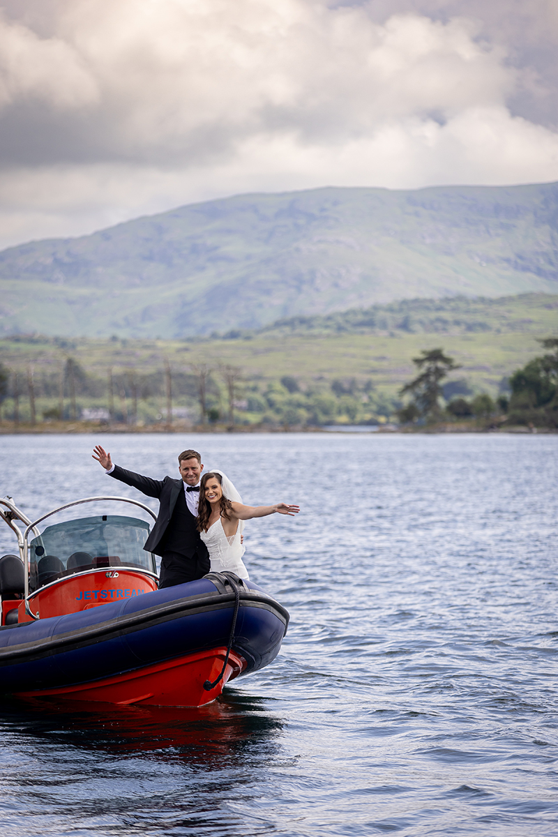 Bride and groom stretch out their arms standing at the front of a boat touring Kenmare bay with a backdrop of green mountains.