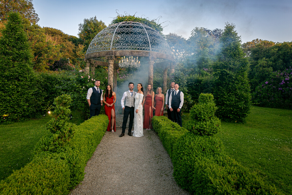 The newlyweds stand in front of a stone gazebo alongside their wedding party in the gardens of Ballyseede castle, surrounded by green foliage and trees.