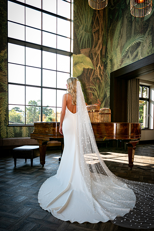 portrait of the bride showcasing the back of her dress standing in front of a grand piano by a window in the dunloe hotel