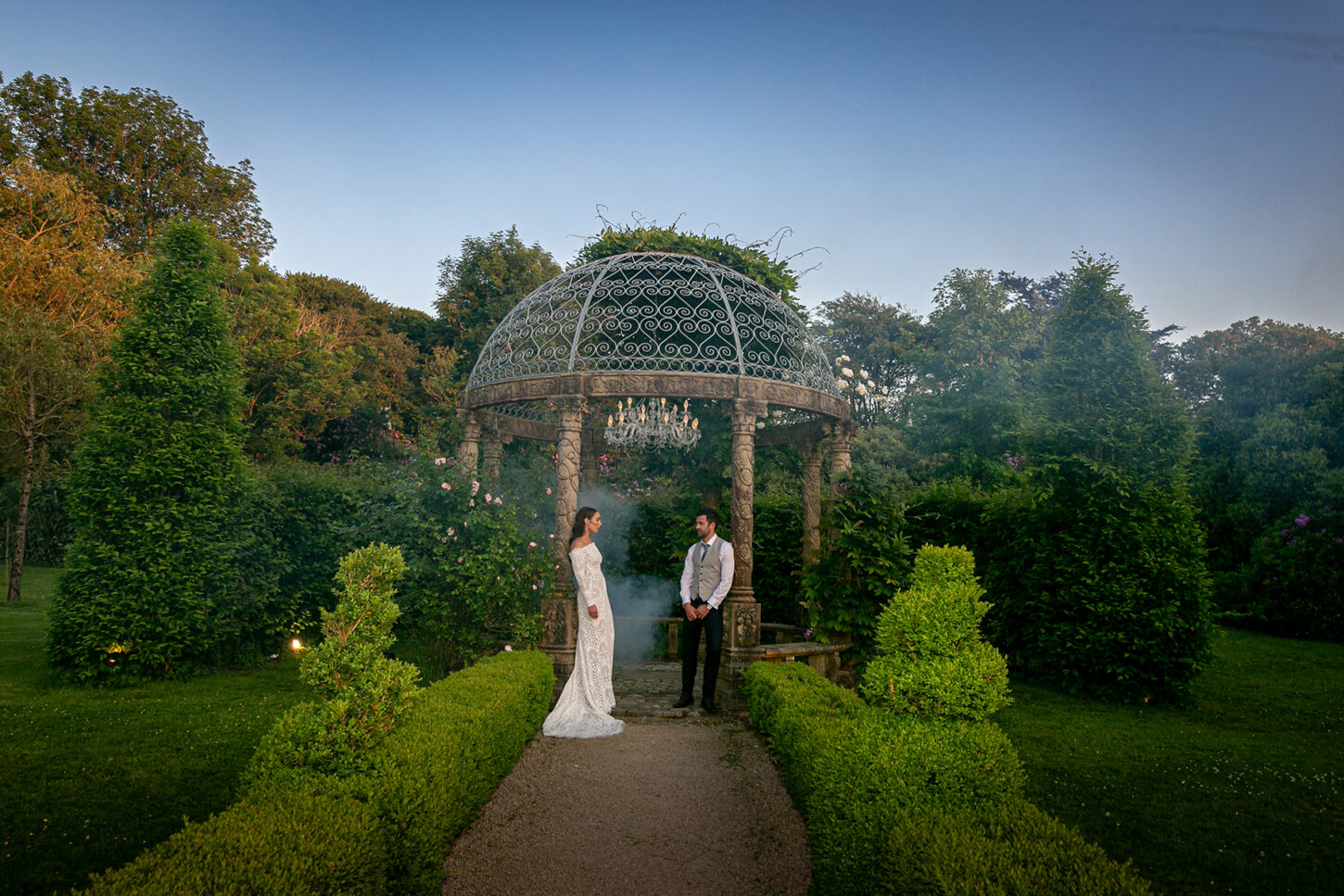Bride and groom lean against the stone gazebo pillars at Ballyseede hotel, Tralee with lush green hedges, shrubs and trees lining the path to the gazebo and visible behind and to the side of the gazebo.