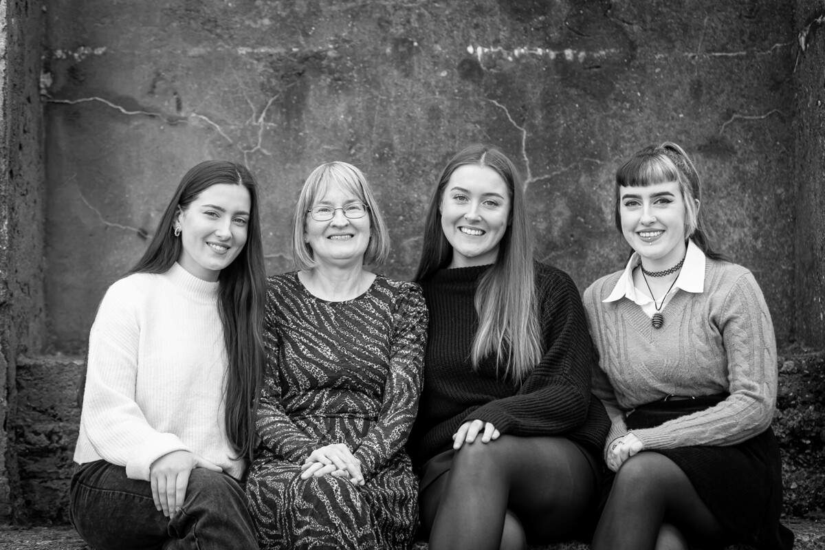 black and white beach portrait of a mother and three daughters sitting against a concrete backdrop