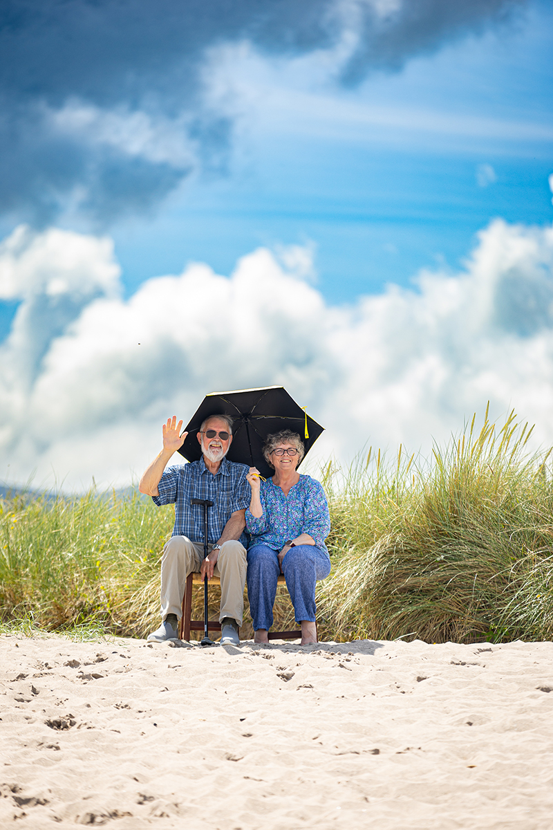 Family beach portrait of grandparents sitting on stools in the sand holding an umbrella with a blue sky and beach dunes in the background.