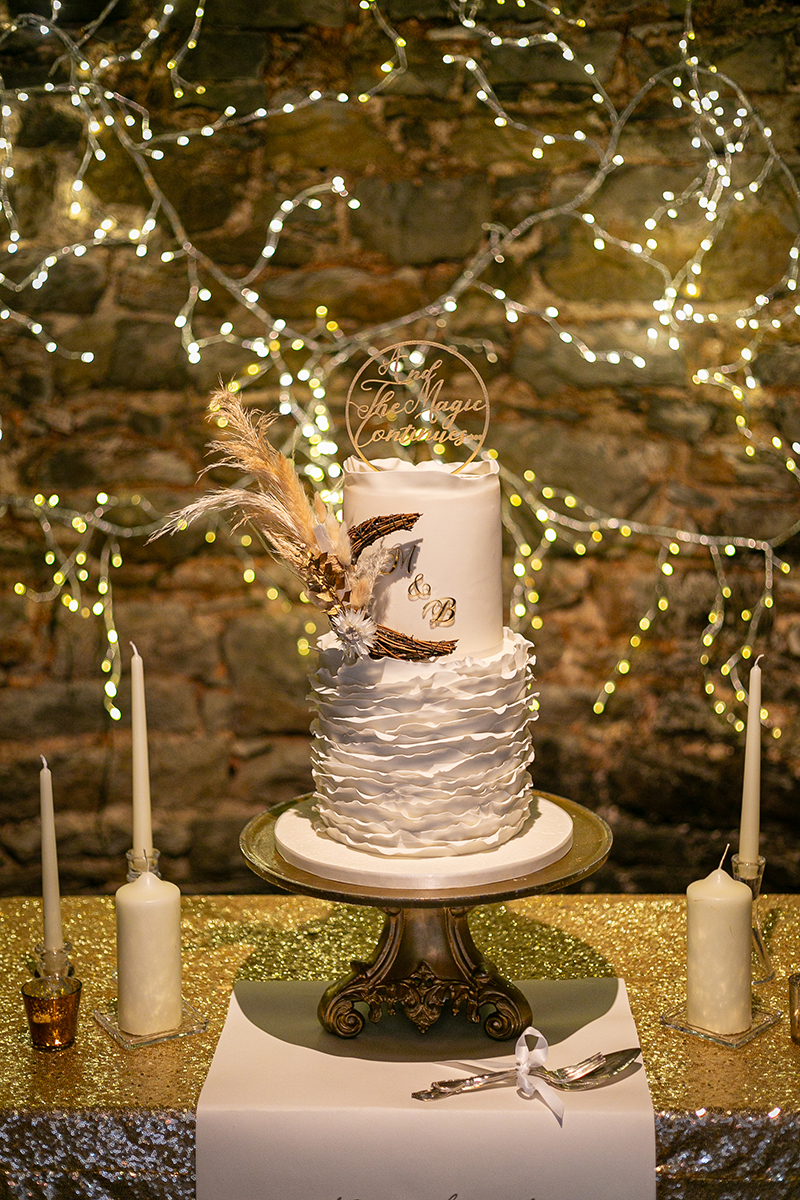A beautiful white wedding anniversary cake for a vow renewal surrounded by gold accents with a stone wall and bokeh lights background at Ballyseede castle, Tralee.