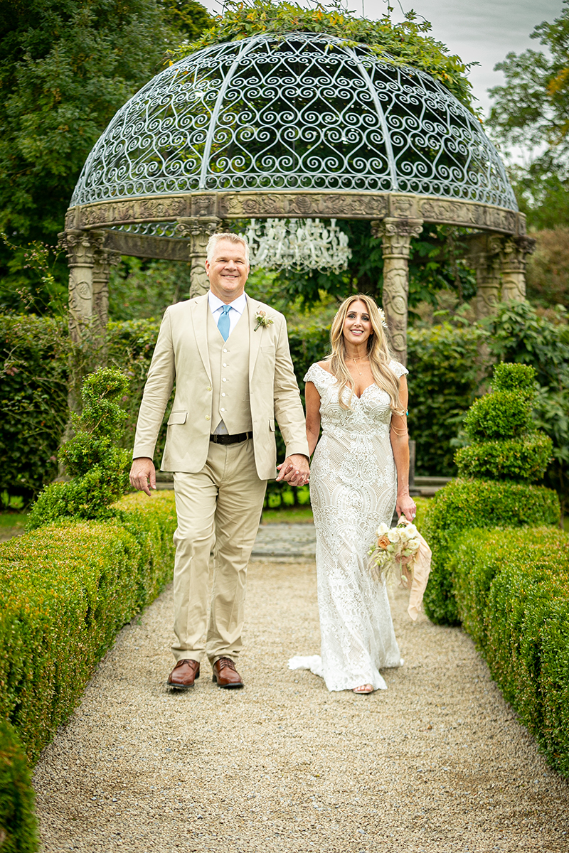 Husband and wife are pictured walking along a path lined by green hedges towards the camera with the stone gazebo, hedges and trees of Ballyseede castle gardens in the background.