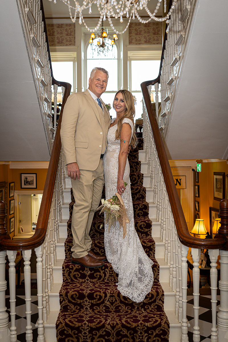 Husband and wife stand on the staircase inside Ballyseede hotel smiling at the camera.