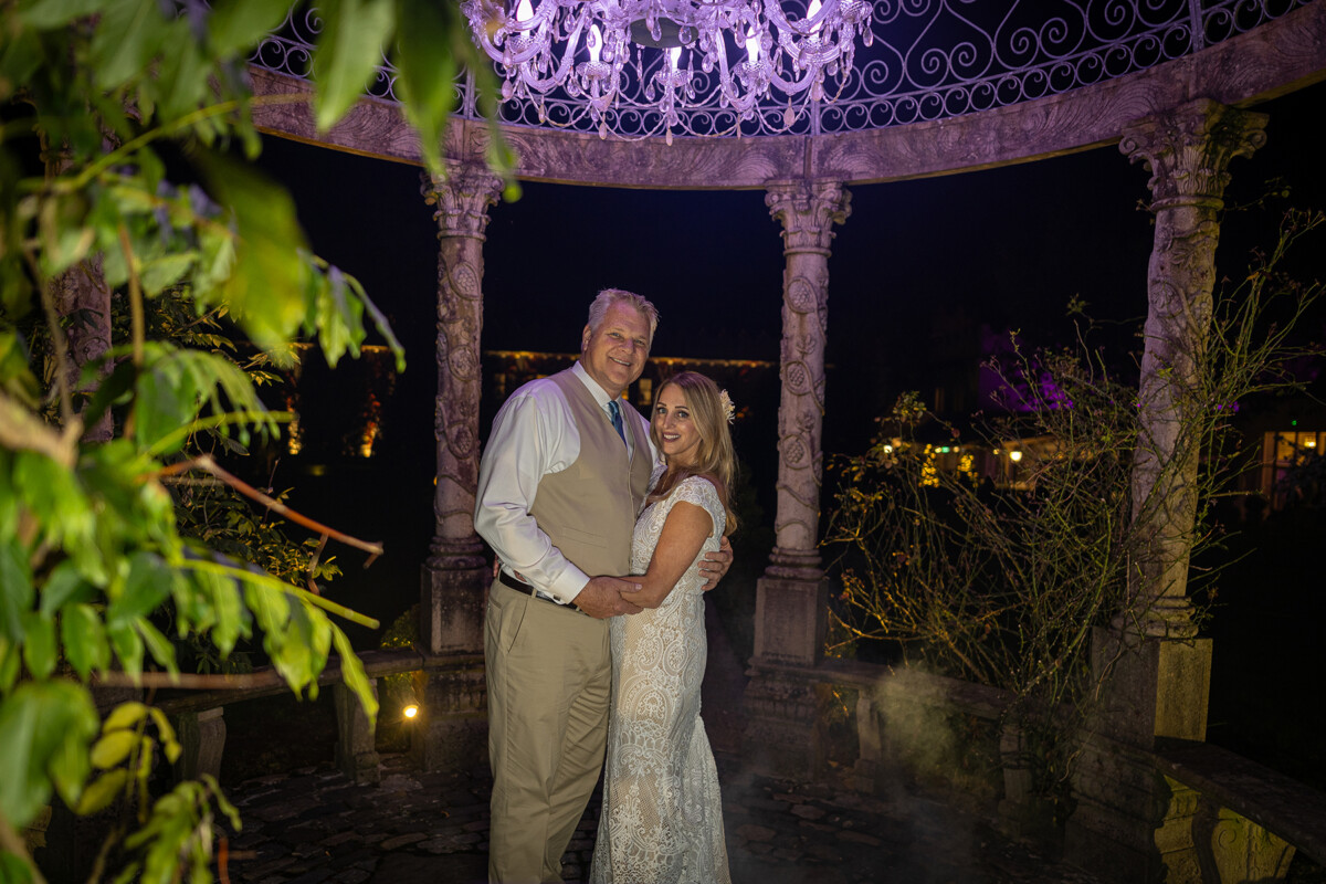 Husband and wife stand arm in arm in the stone gazebo at Ballyseede castle, lit by a chandelier.